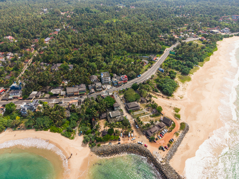 Aerial view of the tropical coastline with a sandy beach coastal road and dense palm forest under bright daylight Peraliya Southern Province Sri Lanka. Aerial view of the tropical coastline with a sandy beach coastal road and dense palm forest under bright daylight Peraliya Southern Province Sri Lanka.
