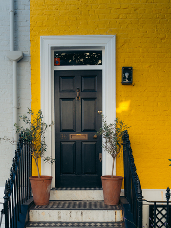 London United Kingdom 21 June 2025 View of a striking black door contrasts against the vibrant yellow facade flanked by potted plants and a white brick wall. London United Kingdom 21 June 2025 View of a striking black door contrasts against the vibrant yellow facade flanked by potted plants and a white brick wall.