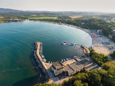Aerial view of the coastal harbor in Kiten with small boats moored along the pier a sandy beach with sun loungers and surrounding lush greenery in Kiten Burgas Bulgaria. Aerial view of the coastal harbor in Kiten with small boats moored along the pier a sandy beach with sun loungers and surrounding lush greenery in Kiten Burgas Bulgaria.