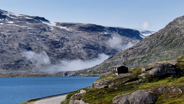Aerial view of a wooden cabin on a rocky outcrop overlooking a blue lake surrounded by rugged mountains with patches of snow and mist in Nordland Norway. Aerial view of a wooden cabin on a rocky outcrop overlooking a blue lake surrounded by rugged mountains with patches of snow and mist in Nordland Norway.