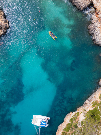 Aerial view of Cala Sa Nau with turquoise waters rocky cliffs a yellow motorboat and a white catamaran in Felanitx Balearic Islands Spain. Aerial view of Cala Sa Nau with turquoise waters rocky cliffs a yellow motorboat and a white catamaran in Felanitx Balearic Islands Spain.