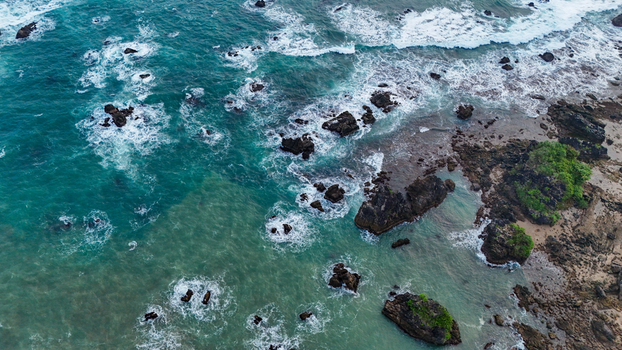 Aerial view of a rocky beach with turquoise waves crashing against dark volcanic rocks and lush green vegetation along the coastline in Indonesia. Aerial view of a rocky beach with turquoise waves crashing against dark volcanic rocks and lush green vegetation along the coastline in Indonesia.
