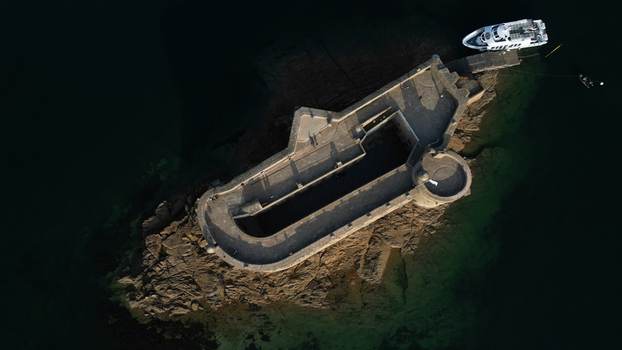 Aerial view of a stark stone fortress bathed in warm sunlight rises from the dark rippling waters a lone boat moored nearby Plouezoc h Bretagne France.