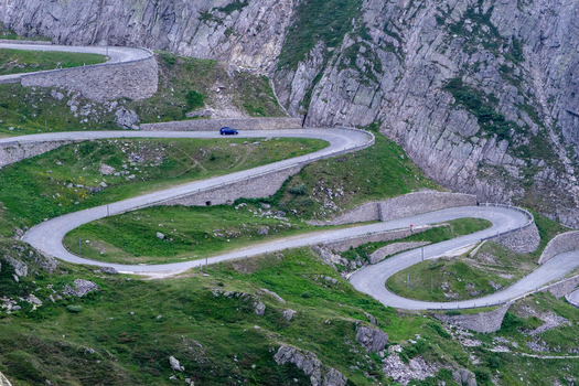 View of the winding Tremola road at Sankt Gotthard Pass with a blue car driving along stone walls and rocky mountain slopes in Airolo Ticino Switzerland.