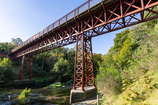 View of The Uncovered Bridge a historic red steel railway bridge spanning a river surrounded by lush green trees under a clear blue sky in Braedstrup Jutland Denmark. View of The Uncovered Bridge a historic red steel railway bridge spanning a river surrounded by lush green trees under a clear blue sky in Braedstrup Jutland Denmark.