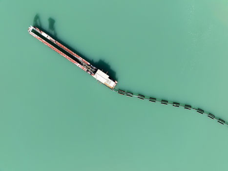 Aerial view of a long industrial barge with a floating containment boom trailing behind on the turquoise water of the Mondego River in Coimbra Coimbra District Portugal. Aerial view of a long industrial barge with a floating containment boom trailing behind on the turquoise water of the Mondego River in Coimbra Coimbra District Portugal.