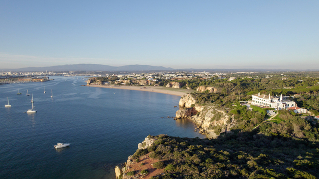 Aerial view of the coastal landscape of Ferragudo featuring cliffs a sandy beach a prominent villa on the headland and boats anchored in the bay in Ferragudo Faro District Portugal.