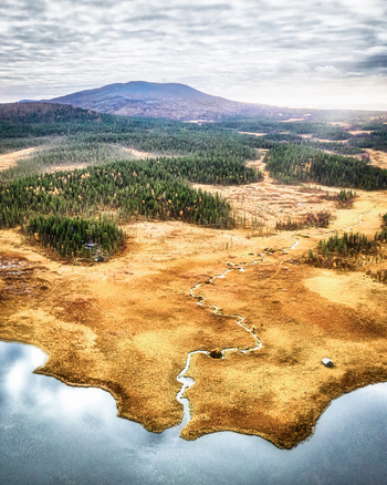 Aerial view of a winding stream flowing through the marshy landscape surrounded by coniferous forests and distant mountains under a cloudy sky in Enontekio Lapland Finland. Aerial view of a winding stream flowing through the marshy landscape surrounded by coniferous forests and distant mountains under a cloudy sky in Enontekio Lapland Finland.