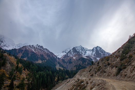 View of rugged snow capped mountain peaks dense pine forests and a winding dirt road under a cloudy sky Naltar Valley Gilgit Pakistan. View of rugged snow capped mountain peaks dense pine forests and a winding dirt road under a cloudy sky Naltar Valley Gilgit Pakistan.