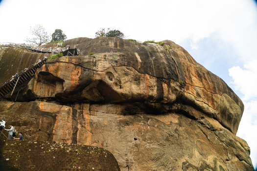 View of the massive Sigiriya Rock Fortress with tourists climbing the steep metal staircases along the ancient granite cliffs in Sigiriya Central Province Sri Lanka. View of the massive Sigiriya Rock Fortress with tourists climbing the steep metal staircases along the ancient granite cliffs in Sigiriya Central Province Sri Lanka.