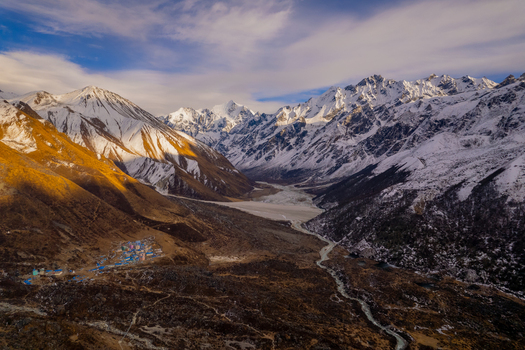 Aerial view of a remote mountain village nestled in a valley surrounded by snow capped Himalayan peaks with a winding river and golden sunlight illuminating the slopes in Nepal.