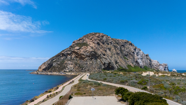 Aerial view of Morro Rock rising above the Pacific Ocean with a coastal road and basketball court in the foreground under a blue sky in Morro Bay California United States.