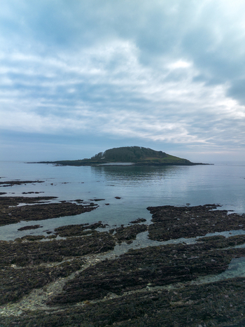 Aerial view of Looe Island surrounded by calm sea with rocky shoreline in the foreground under a cloudy sky in Looe Cornwall United Kingdom. Aerial view of Looe Island surrounded by calm sea with rocky shoreline in the foreground under a cloudy sky in Looe Cornwall United Kingdom.
