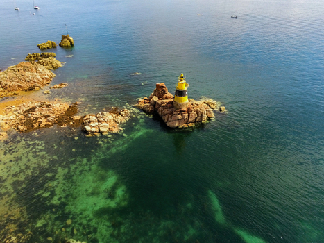 Aerial view of the yellow and black lighthouse standing on a rocky outcrop surrounded by clear turquoise and deep blue sea waters in Ile de Brehat Brittany France.
