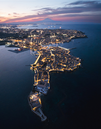 Aerial view of the illuminated Island of Ortigia at dusk with Mount Etna visible in the distance under a twilight sky in Syracuse Sicily Italy.