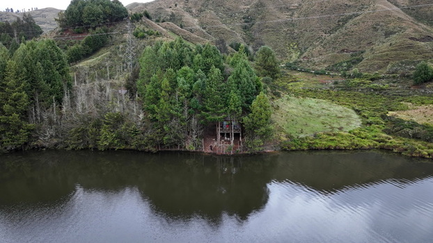 Aerial view of Corani Lake with a small wooden structure nestled among dense pine trees on the shoreline surrounded by rolling hills in Chapare Cochabamba Department Bolivia. Aerial view of Corani Lake with a small wooden structure nestled among dense pine trees on the shoreline surrounded by rolling hills in Chapare Cochabamba Department Bolivia.