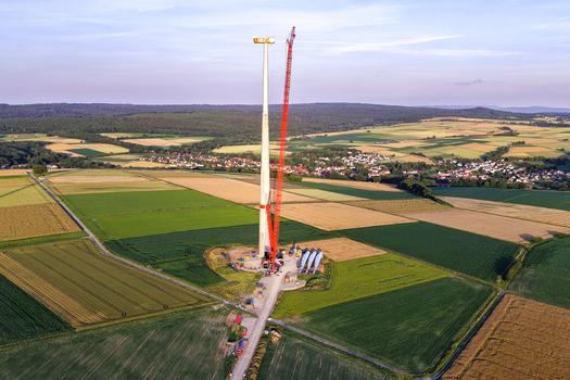 Aerial view of a wind turbine construction site featuring a tall crane turbine tower and blades resting on the ground amidst agricultural fields in Hofgeismar Hessen Germany.