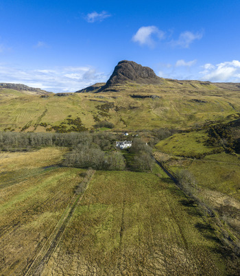 Aerial view of a white house nestled in a valley below a prominent rocky hill and rolling green fields Isle of Skye Scotland United Kingdom. Aerial view of a white house nestled in a valley below a prominent rocky hill and rolling green fields Isle of Skye Scotland United Kingdom.