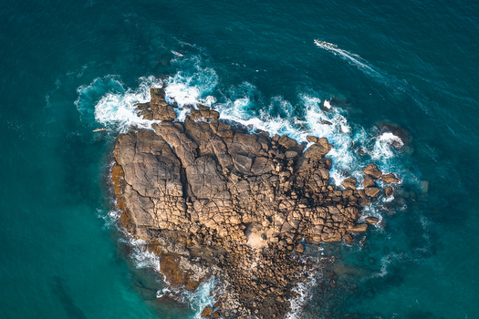 Aerial view of a large rocky island surrounded by turquoise ocean water and crashing waves with a small boat nearby Kovalam Kerala India. Aerial view of a large rocky island surrounded by turquoise ocean water and crashing waves with a small boat nearby Kovalam Kerala India.