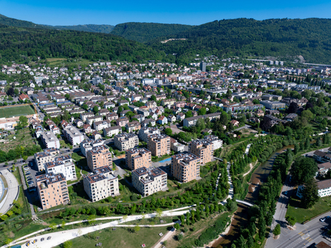 Aerial view of modern residential apartment blocks nestled between a winding river and lush forested hills under a clear blue sky in Biel Bienne Canton of Bern Switzerland.