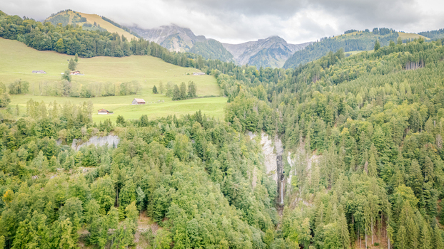Aerial view of Grandvillard Waterfall cascading through a dense green forest with alpine meadows and mountain peaks under a cloudy sky in Grandvillard Fribourg Switzerland.