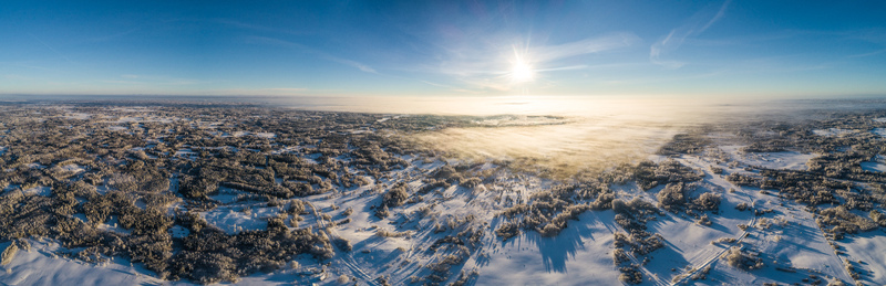 Aerial view of a vast snow covered landscape in Latvia featuring dense forests open fields and a layer of low lying morning mist illuminated by the bright sun. Aerial view of a vast snow covered landscape in Latvia featuring dense forests open fields and a layer of low lying morning mist illuminated by the bright sun.