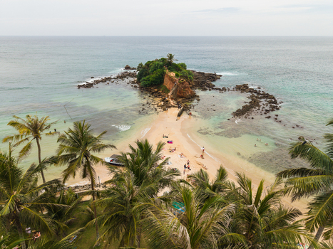 Aerial view of Parrot Rock and the sandy beach with people sunbathing by turquoise waters and palm trees in Mirissa Southern Province Sri Lanka. Aerial view of Parrot Rock and the sandy beach with people sunbathing by turquoise waters and palm trees in Mirissa Southern Province Sri Lanka.