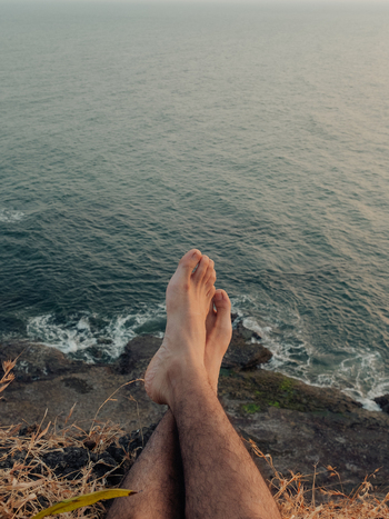 View of bare feet dangling over a rugged cliff edge where golden grass meets the turquoise sea in a serene coastal moment Chiplun Maharashtra India. View of bare feet dangling over a rugged cliff edge where golden grass meets the turquoise sea in a serene coastal moment Chiplun Maharashtra India.
