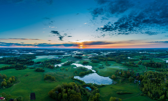Aerial view of a serene landscape featuring lush green fields small reflective ponds and patches of forest under a dramatic sunset sky in Latvia. Aerial view of a serene landscape featuring lush green fields small reflective ponds and patches of forest under a dramatic sunset sky in Latvia.