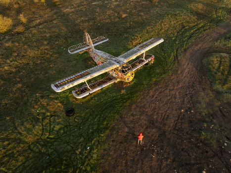 Aerial view of an abandoned biplane with graffiti on its fuselage resting in a grassy field with tire tracks and a person in a red jacket standing nearby in Bulgaria. Aerial view of an abandoned biplane with graffiti on its fuselage resting in a grassy field with tire tracks and a person in a red jacket standing nearby in Bulgaria.