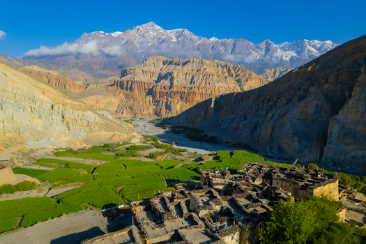 Aerial view of a traditional village with stone houses and vibrant green terraced fields nestled in a deep valley surrounded by rugged Himalayan mountains in Nepal.