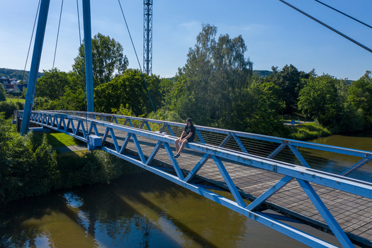 Aerial view of the blue metal bridge at Sportinsel Forchheim with a person sitting on the railing over the river surrounded by green trees in Forchheim Bayern Germany.