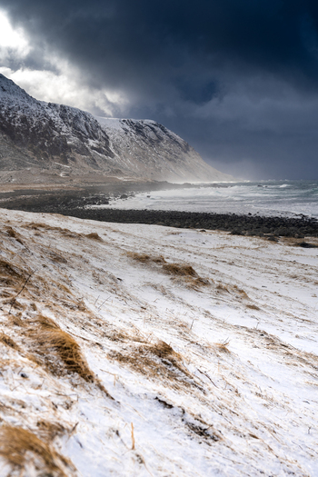 View of a snow covered beach and rugged mountains under a dramatic dark stormy sky in Leknes Lofoten Norway.