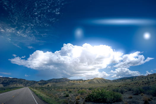 View of a road leading towards the landscape of Bryce Canyon National Park under a bright blue sky with a large cumulus cloud and lens flare in Utah United States.