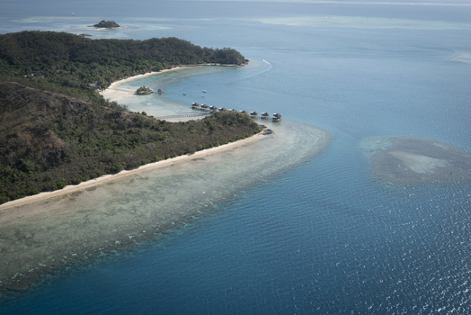 View of Likuliku Lagoon Resort with overwater bungalows sandy beach and turquoise lagoon surrounded by lush green hills Yaro Nadroga Navosa Fiji.