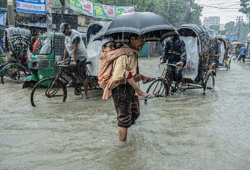 Chattogram Bangladesh 28 July 2025 View of a man carrying a child on his back through a flooded street during heavy rain with rickshaws and people navigating the water.