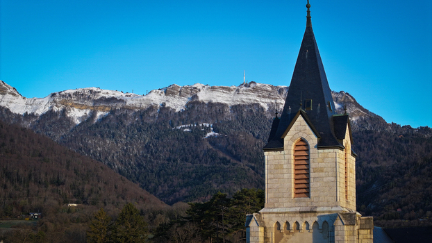 Aerial view of the stone tower of Eglise Saint Pierre with snow capped mountains and a clear blue sky in the background Gex Auvergne Rhone Alpes France.