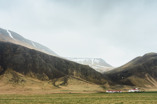 View of a small white house with a red roof nestled at the base of rugged snow dusted mountains under a cloudy sky in the Icelandic countryside Reykjavik Reykjavikurborg Iceland.