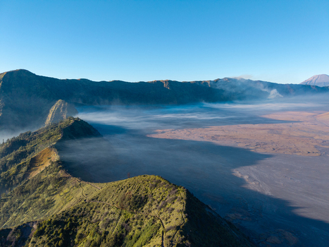 Aerial view of the volcanic landscape of Mount Bromo with mist rolling over the vast caldera and rugged mountain ridges under a clear blue sky in Sukapura East Java Indonesia. Aerial view of the volcanic landscape of Mount Bromo with mist rolling over the vast caldera and rugged mountain ridges under a clear blue sky in Sukapura East Java Indonesia.