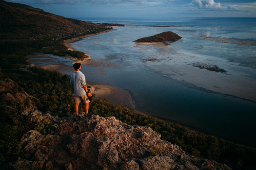 Aerial view of the coast of Mauritius Island with a man standing on a rocky cliff overlooking a turquoise lagoon and a small island at sunset Mauritius. Aerial view of the coast of Mauritius Island with a man standing on a rocky cliff overlooking a turquoise lagoon and a small island at sunset Mauritius.