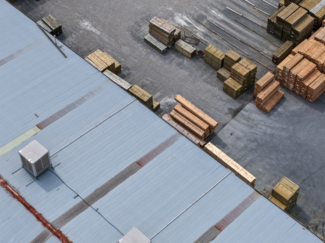 Aerial view of an industrial site with stacks of timber and lumber stored on concrete ground next to a large warehouse roof in Melbourne VIC Australia.