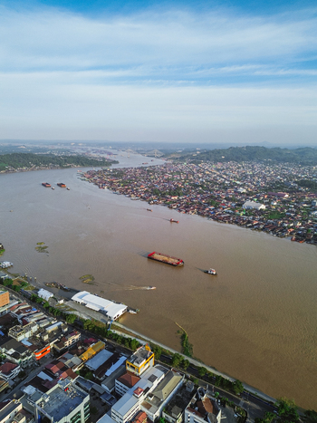 Aerial view of the Mahakam River flowing through the city of Samarinda with a barge navigating the brown water urban rooftops and lush green hills in Samarinda Kalimantan Timur Indonesia. Aerial view of the Mahakam River flowing through the city of Samarinda with a barge navigating the brown water urban rooftops and lush green hills in Samarinda Kalimantan Timur Indonesia.