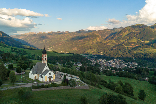 Aerial view of Fiemme Valley featuring a historic church with a bell tower and cemetery surrounded by lush green meadows and mountains in Trentino Alto Adige Italy.