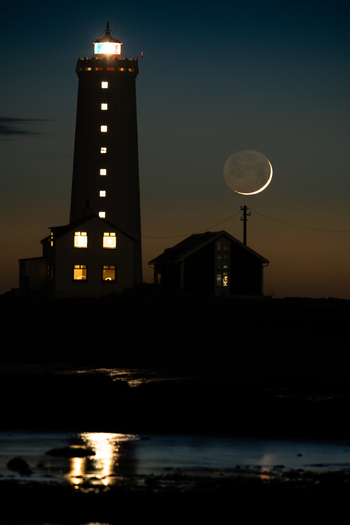 View of Grotta Lighthouse at night with a crescent moon and glowing windows in nearby houses Seltjarnarnes Seltjarnarnesbaer Iceland. View of Grotta Lighthouse at night with a crescent moon and glowing windows in nearby houses Seltjarnarnes Seltjarnarnesbaer Iceland.