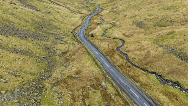 Aerial view of a winding road cutting through the rugged grassy landscape contrasting with the dark asphalt and the meandering stream England United Kingdom. Aerial view of a winding road cutting through the rugged grassy landscape contrasting with the dark asphalt and the meandering stream England United Kingdom.