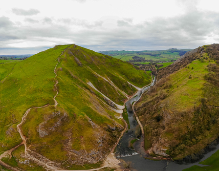 Aerial view of Dovedale featuring the winding River Dove lush green hills and walking paths under a cloudy sky in Ashbourne England United Kingdom. Aerial view of Dovedale featuring the winding River Dove lush green hills and walking paths under a cloudy sky in Ashbourne England United Kingdom.