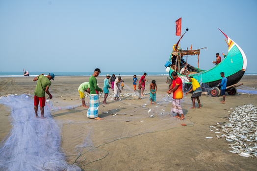 Teknaf Bangladesh 10 March 2026 View of fishermen casting nets and sorting their catch on the sandy beach their vibrant clothing contrasting with the muted tones of the shore.