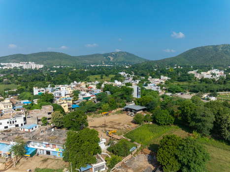 Aerial view of a residential area in Udaipur Rajasthan India featuring scattered buildings lush green trees and rolling hills in the background under a clear blue sky. Aerial view of a residential area in Udaipur Rajasthan India featuring scattered buildings lush green trees and rolling hills in the background under a clear blue sky.