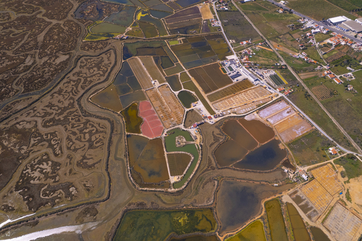 Aerial view of the Salinas do Grelha salt pans featuring geometric ponds with varying water colors surrounded by marshlands and coastal terrain in Olhao Faro Portugal. Aerial view of the Salinas do Grelha salt pans featuring geometric ponds with varying water colors surrounded by marshlands and coastal terrain in Olhao Faro Portugal.