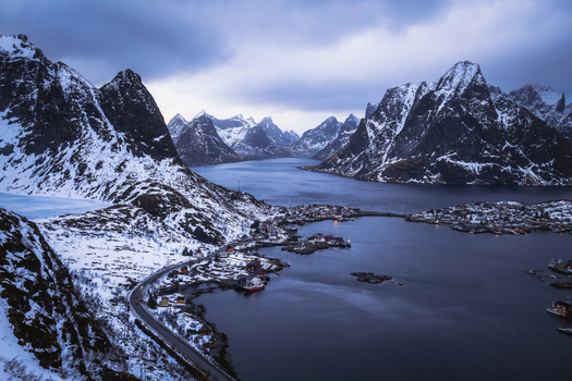 Aerial view of dramatic snow dusted peaks embracing the still dark waters dotted with tiny vibrant villages under a brooding sky Reinebringen Nordland Norway.
